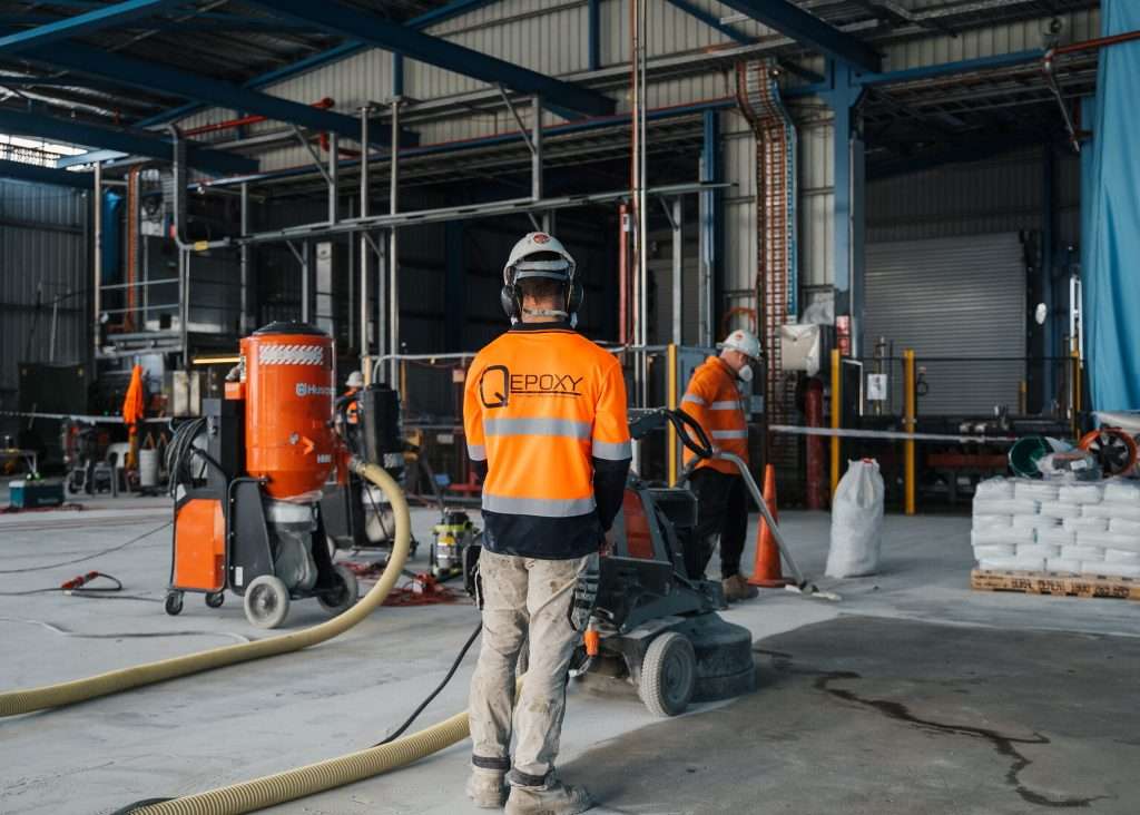 Two Qepoxy operators running floor grinders and a dust extractor across a bare concrete slab
